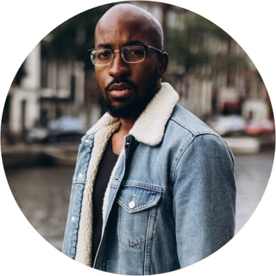 Dave Harris has dark skin, deep brown eyes, black facial hair, and is bald. He wears half-framed glasses and a denim jacket with a sherpa collar over a black tee.