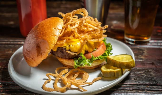 A cheeseburger with the top bun falling off. Fried onions are stacked on top and topple over. There are two pickle slices on the side.