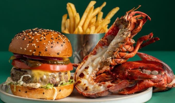 A burger and a half lobster tail and a cup of fries on a white plate in front of a green background. 