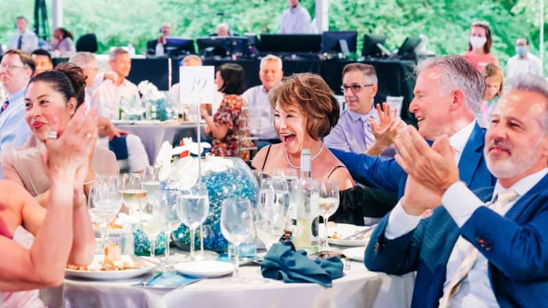 A group of smiling people wearing formal attire clap while sitting at formal dining tables under an outdoor pavillion.