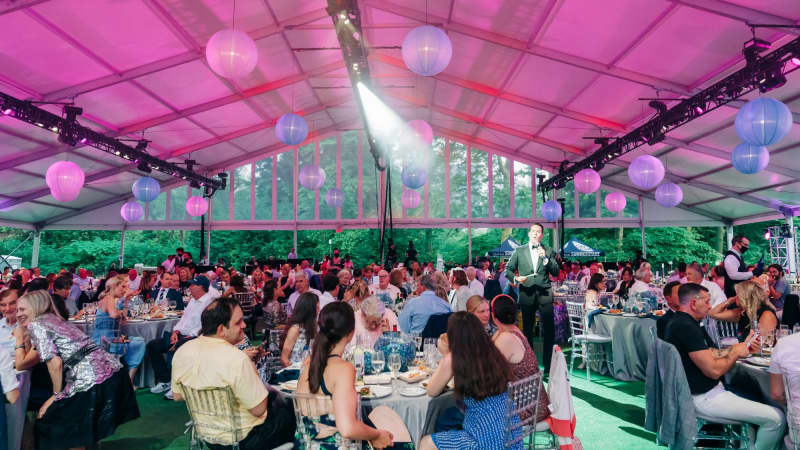 A wideshot of a large outdoor pavillion with pink lighting. Numerous round dining tables fill the room and people in formal attire sit at them.