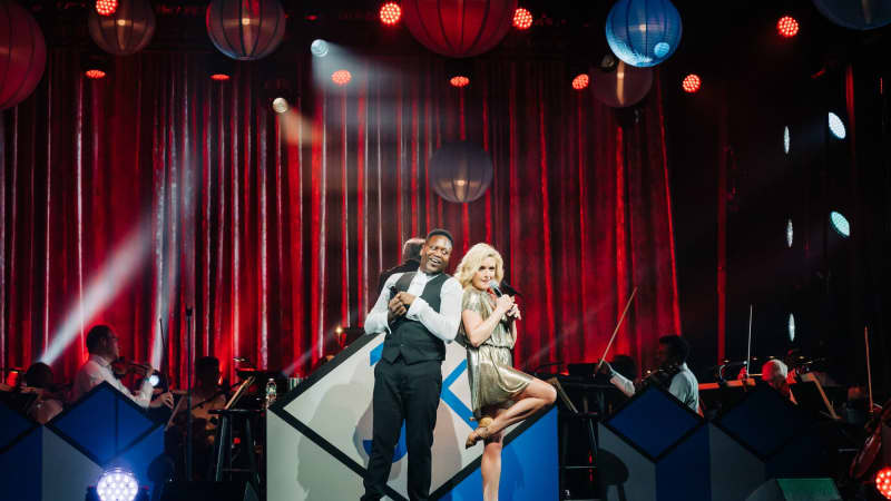 Tituss Burgess and Jane Krakowski perform together on a stage with a red-lit curtain behind them.