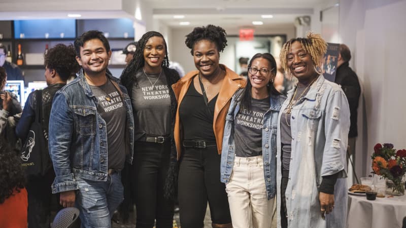 Five people standing together with their arms around each other smiling and posing for a picture. All five of them are wearing New York Theatre Workshop. 