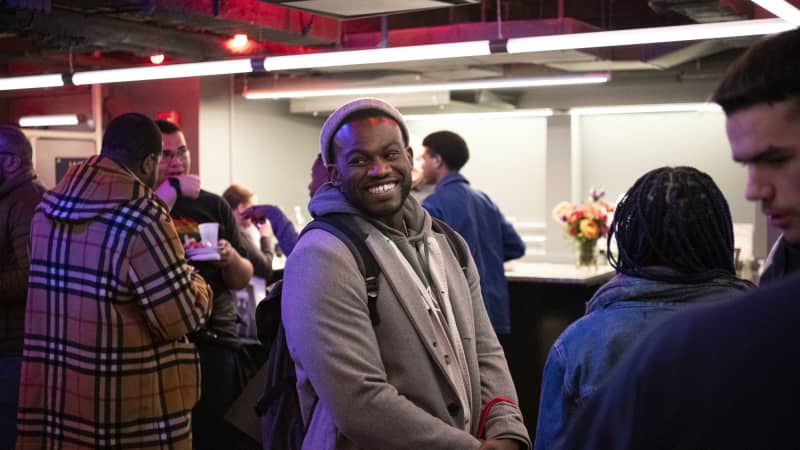 A dark skinned man standing in a busy room smiling at the camera wearing a grey sweater and a black backpack. 