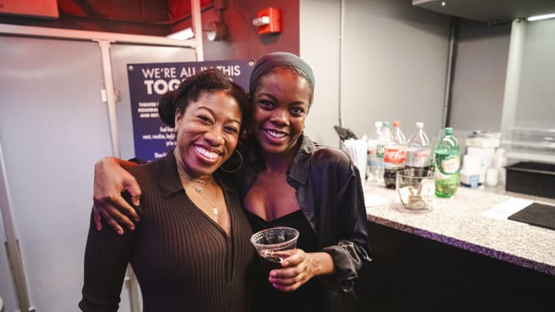 Two dark skinned girls wearing black smiling at the camera with their arms around one another. 