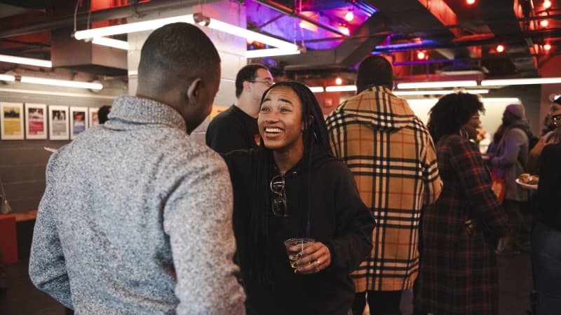 Two dark skinned individuals, one woman and one man, talking with eahcother. The woman is facing the camera and smiling while we only see the back of the short haired man in a gray sweater. 