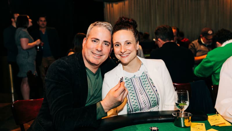 A man and woman sitting at a green poker table lean in close and smile. The man holds up a few poker chips in his hand.