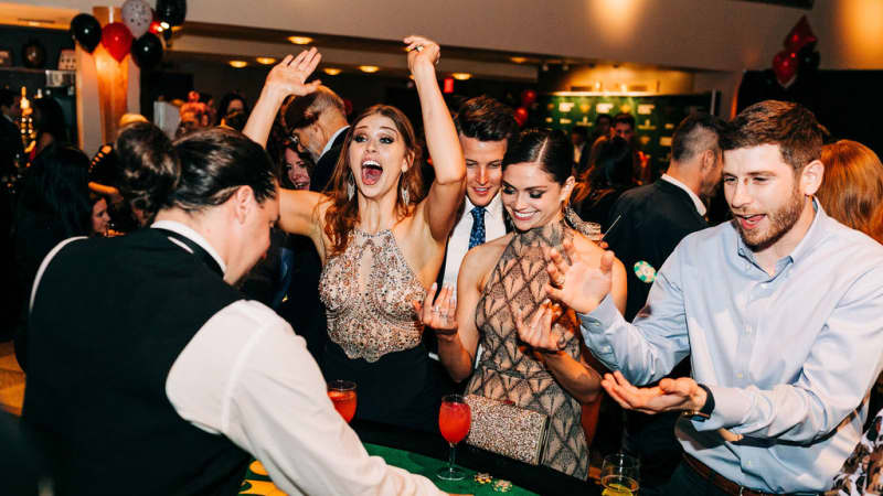 A group of people at a green casino table cheer and clap as the dealer places cards on the table.