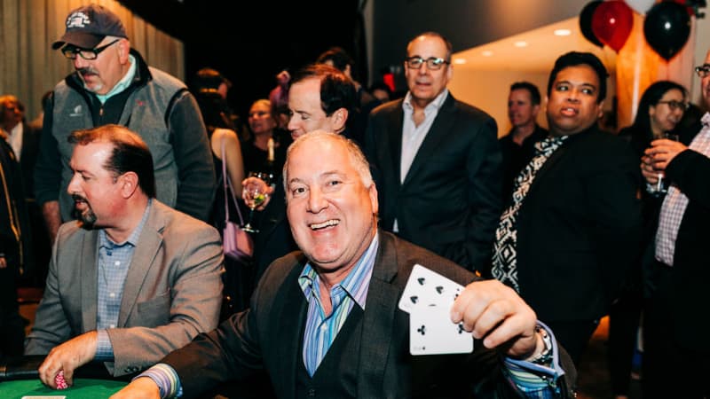 An older man in a suit sits at a small green casino table and holds up two playing cards to the camera.