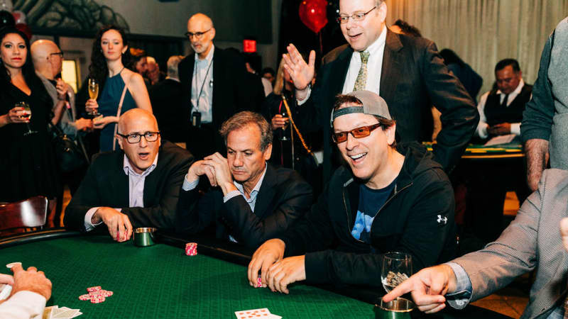 A line of men sit at a green casino table with poker chips in their hands as they watch the dealer shuffle cards.