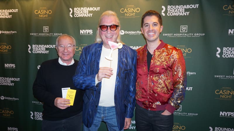 Three men stand in front of a green step and repeat. One of them holds up a prop paper margarita glass on a stick and wears sunglasses.