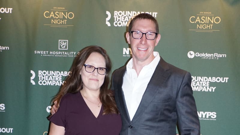 A woman in a dark purple jump suit and glasses puts her arm around a man in a gray suit with glasses for a photo in front of a green step and repeat.