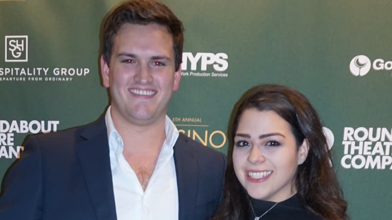 A man in a navy suit and white collared shirt poses with a woman with long dark hair in a black top and pants. They both hold glasses of liquor.