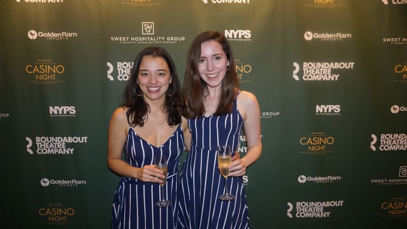 Two women wearing matching blue and navy vertical-striped dresses hold champagne glasses and pose together for a photo in front of a green step and repeat.