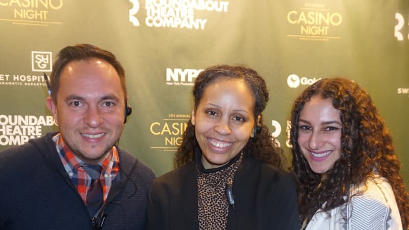 A man and two women pose together for a photo in front of a green step and repeat paterned with several logos.