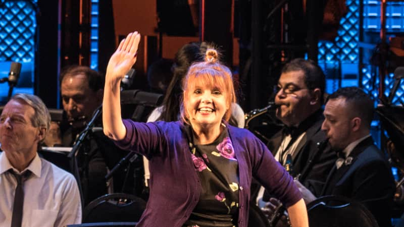 Annie Golden waves on stage in front of an orchestra. She wears a purple cardigan over a black dress with purple roses.