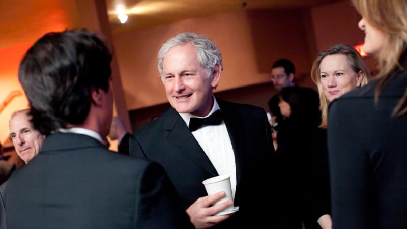 Victor Garber wearing a tuxedo holds a cup and talks to a group of people.