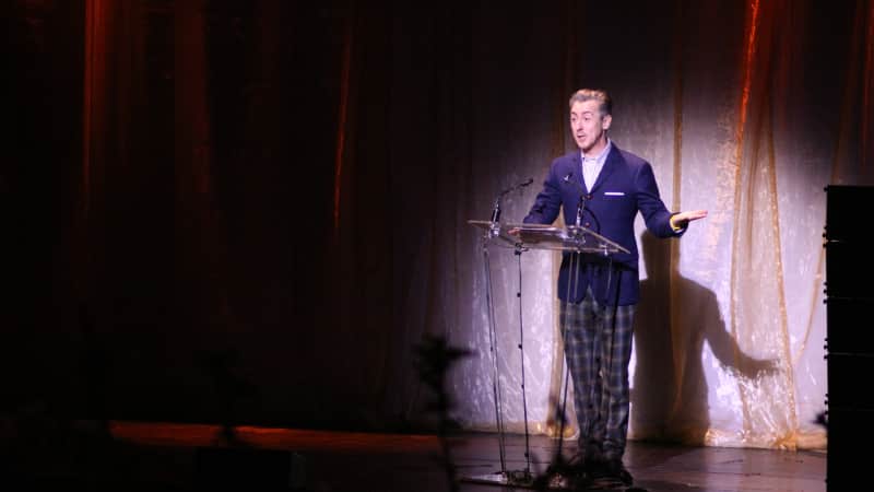 Alan Cumming stands on stage at a glass podium illuminated by a singular spotlight.