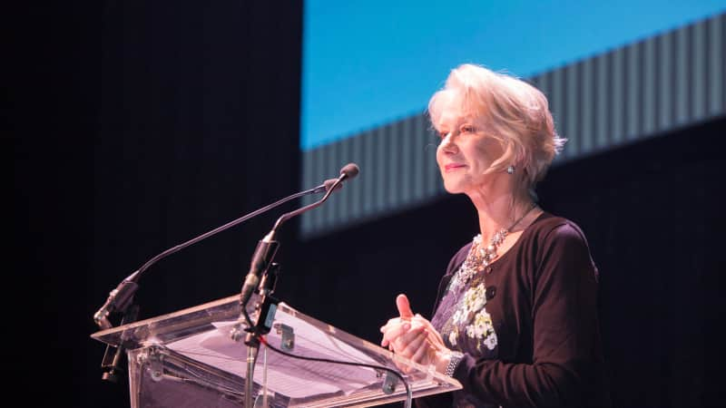 A woman with white hair speaks behind a glass podium on a stage.