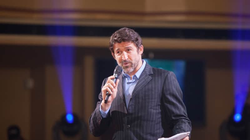 A man with dark brown hair wearing a suit speaks into a microphone and holds a piece of paper.