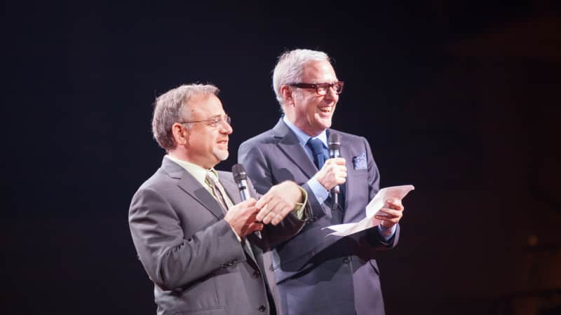 Two men in suits stand on stage and speak into microphones.