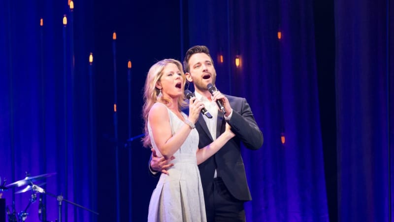 Kelli O'Hara and Colin Donnell hold each other and sing into hand-held mics in front of a blue background with amber sparkles on it.