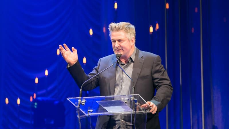 Alec Baldwin speaks at a glass podium on stage in front of a blue stage background.