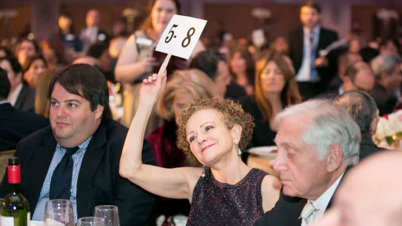 A woman in a crowded ballroom holds up an auction sign while sitting at a fancy dining table.