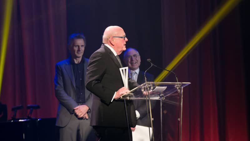 Board Chairman Tom Tuft, an older man wearing a suit, stands at a podium holding an award.