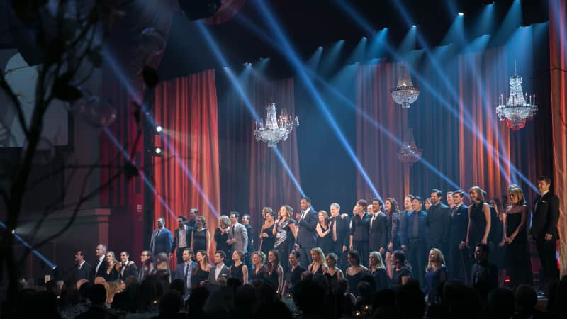 A choir of two rows of people dressed in black perform on a stage with red curtains. Streaks of blue lights shine on them.