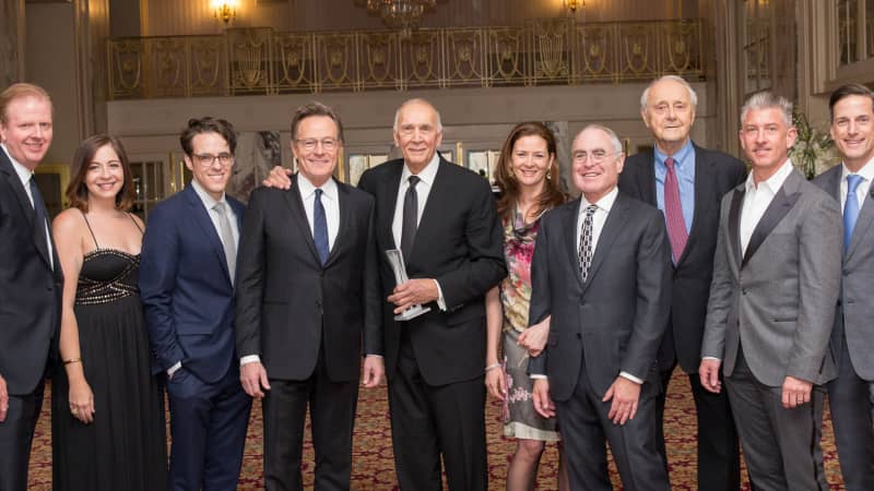 A group of eleven people in formal attire stand in a line and smile for a photo.