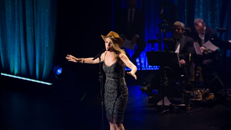 A blonde woman in a black sparkly dress wears a brown cowboy hat and sings on stage.