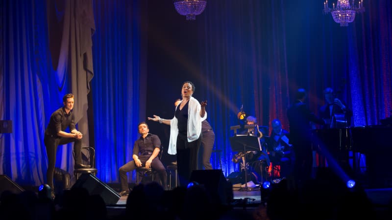 A woman in a black dress and white shawl sings on stage. Behind her is a band and a few men dressed in black lounging on cabaret chairs.