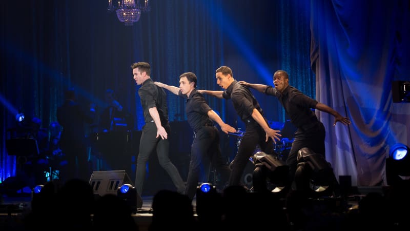 Four men dressed in black dance on stage. They stand in a line behind one another, resting one arm on the shoulder in front of them and swinging the other by their sides.