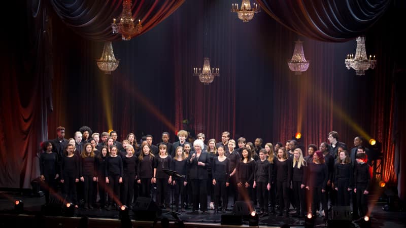 A large choir of people dressed in black sing on stage under several chandeliers.