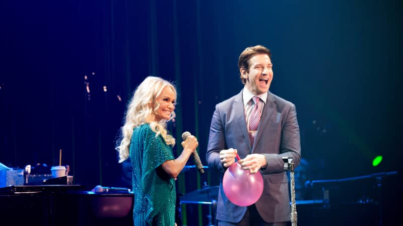 Kristin Chenoweth holds a microphone and smiles while Andy Karl laughs and holds a balloon on stage.