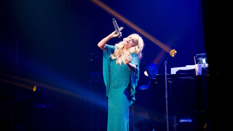 Kristin Chenoweth wears a long sparkly turquoise dress and sings passionately into a microphone.