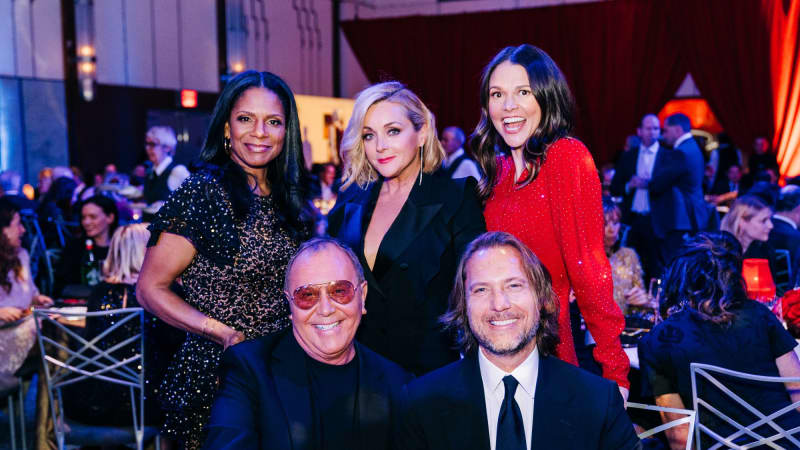 Audra McDonald, Jane Krakowski, and Sutton Foster smile while standing behind seated Michael Kors and Lance LePere at their dining table.