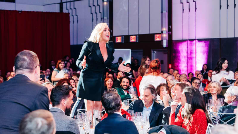 In a crowded Ziegfeld Ballroom full of people in formal attire sitting at dining tables, Jane Krakowski stands up on her chair at her table and speaks to the room.