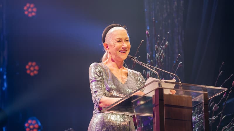 Helen Mirren, an older woman with white hair in a ponytail, wears a silver dress and reads a speech behind a glass podium.