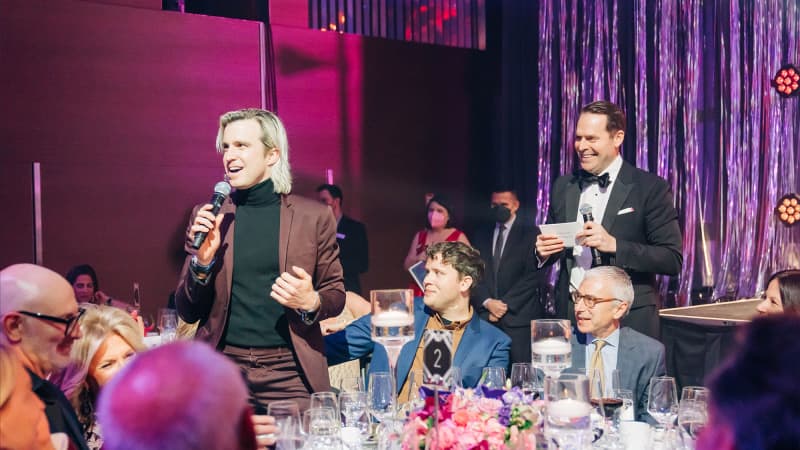 Two people stand and hold micrphones in a room full of people sitting at fancy dining tables under a wash of pink and purple lighting.