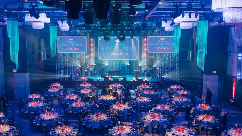 A wideshot of the Ziegfeld Ballroom for the 2022 gala. Numerous round dining tables are scattered in a room washed with blue lighting. A stage is at the front of the room.