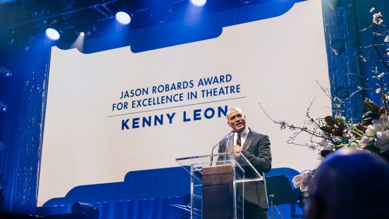 Kenny Leon with dark skin and bald head wearing a suit giving a speech at a podium on a stage. 