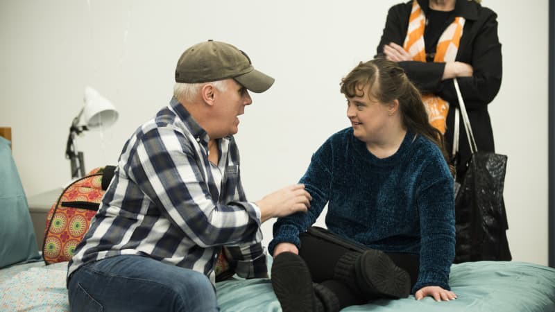 Director Scott Ellis sits next to actress Jamie Brewer on a bed in the rehearsal studio for AMY AND THE ORPHANS.