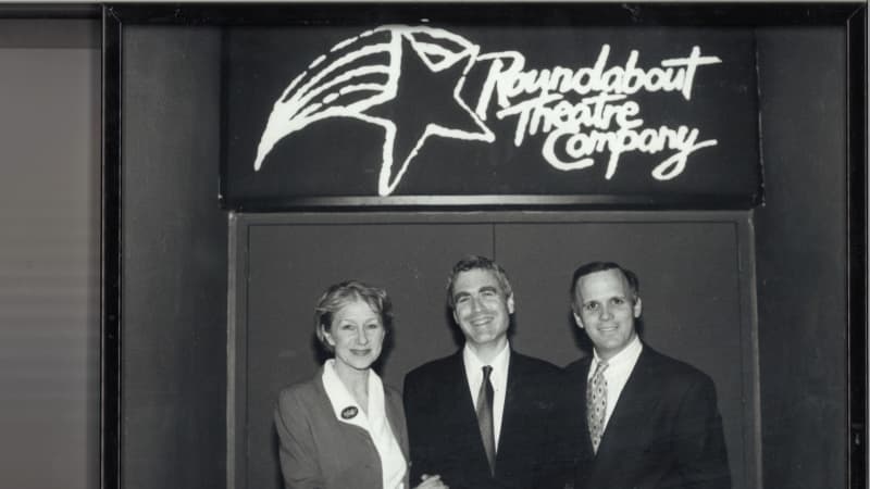 A black and white photo of Helen Mirren, Todd Haimes, and Scott Ellis standing below an old glowing Roundabout sign.