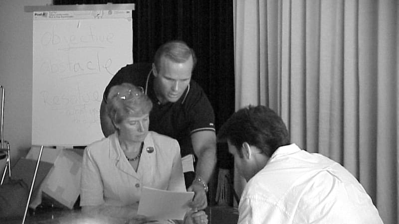 A black and white photo of two actors sitting across from each other holding scripts. A man leans over the woman's shoulder and puts a hand on her script.