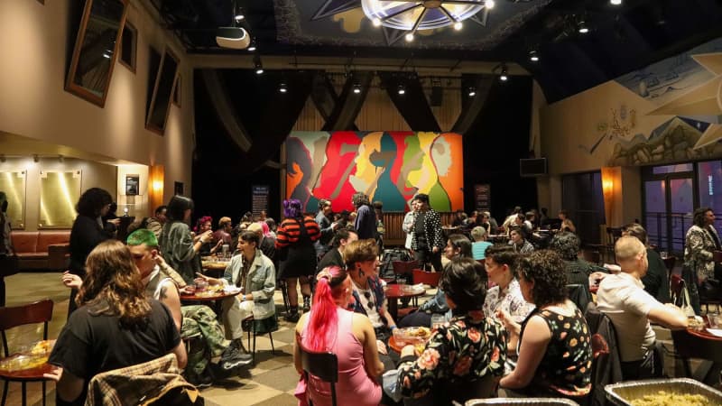 A large group of people eat food at tables in a spacious, decadent lobby. There are trays of catered food lined up. On one wall is a large 1776 mural with colorful side profiles on it.