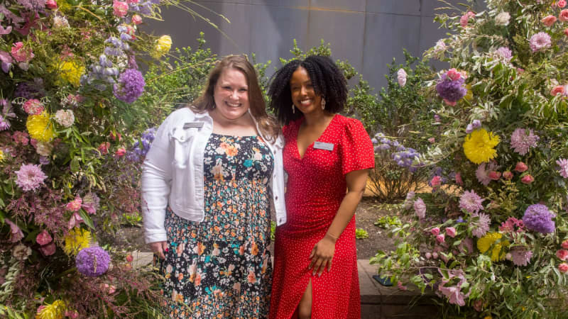 Two women with name tags smile together of a photo near large, colorful floral arrangements.