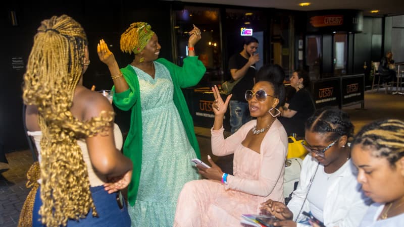 A group of five women, some sitting and some standing, talk and smile outside while gesturing their arms in the air. One woman has her hair in a headwrap and another has long golden braids.