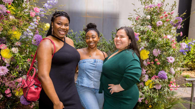 Three women put their arms around each others' waists and pose for a photo near two large floral arrangements.